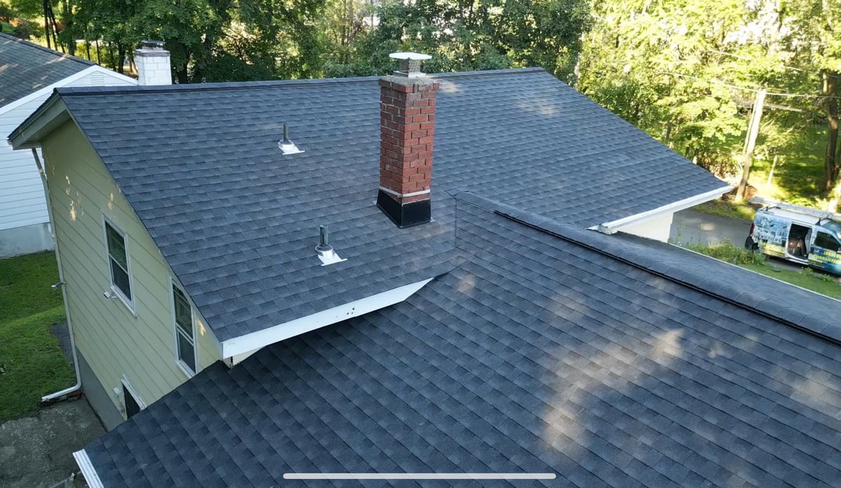 Aerial view of a residential house roof with dark asphalt shingles, brick chimney, and white gutters surrounded by green trees