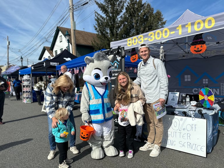 Family posing with a costumed mascot character at an outdoor community event with vendor tents and Halloween decorations
