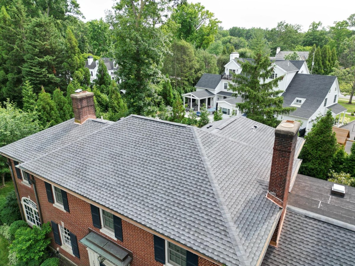 Aerial view of brick house with gray shingle roof and chimneys, surrounded by green trees and residential neighborhood