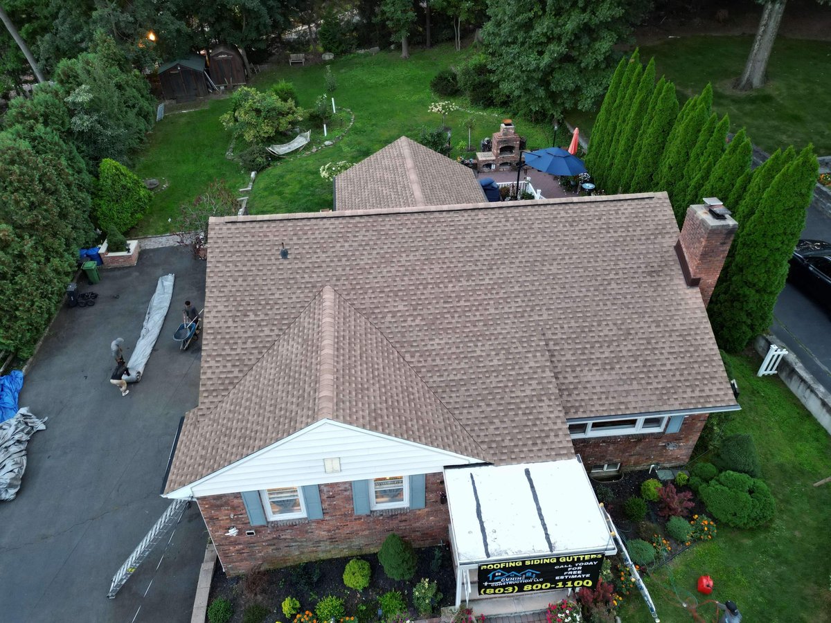 Aerial view of brick house with gray roof and attached carport surrounded by green lawn and trees