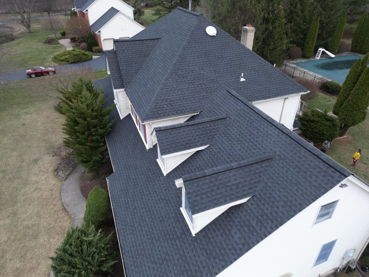 Aerial view of a residential home with dark gray shingle roof, white trim, and multiple skylights surrounded by landscaping