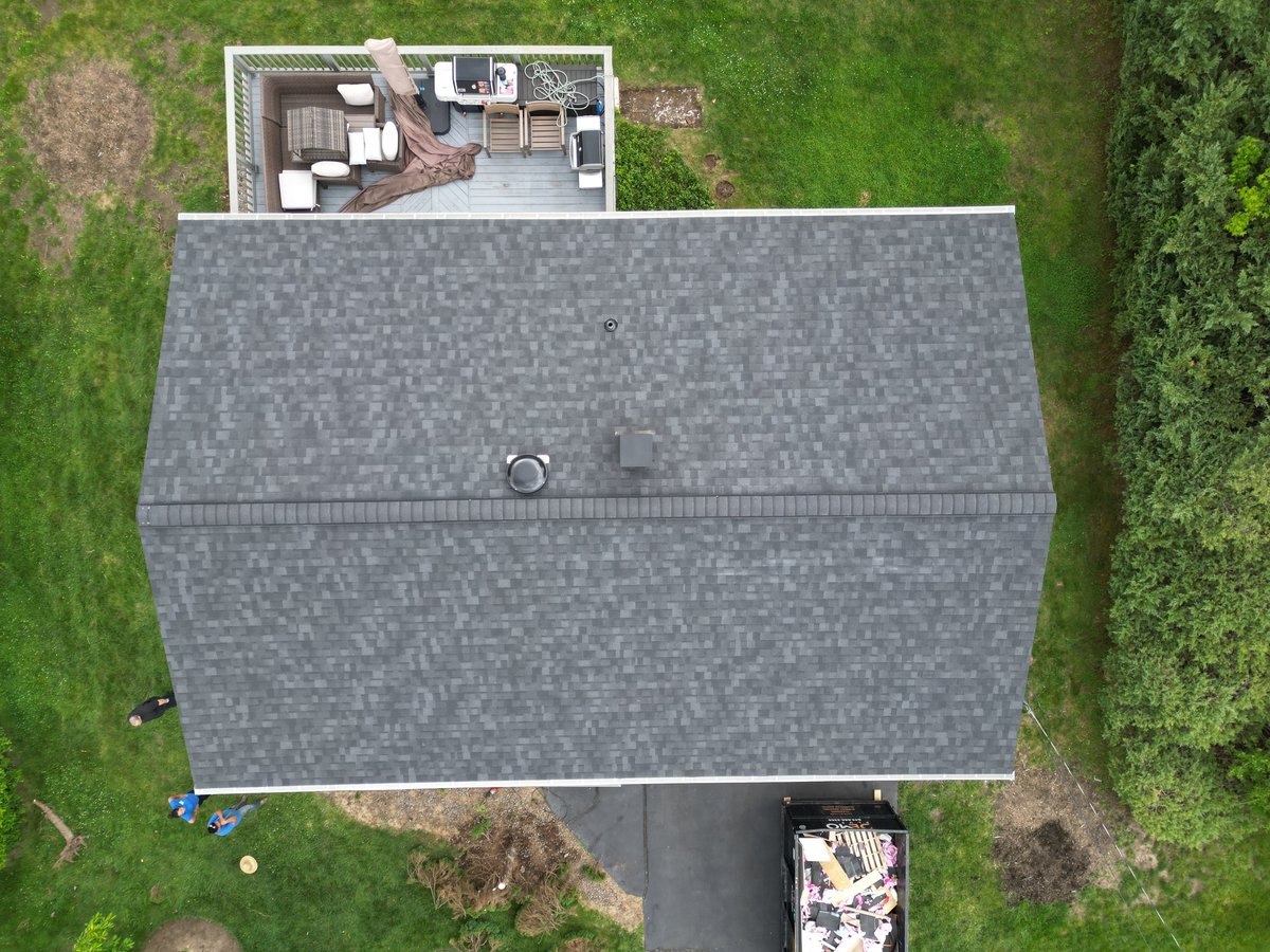 Aerial view of a house with gray asphalt shingle roof, driveway, and surrounding green lawn and trees